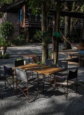 Eski ahşap masa ve evin önünde katlanan boş kamp sandalyeleri. Sabah güneşinin ışıldadığı doğa ortamı. Seçici odak.