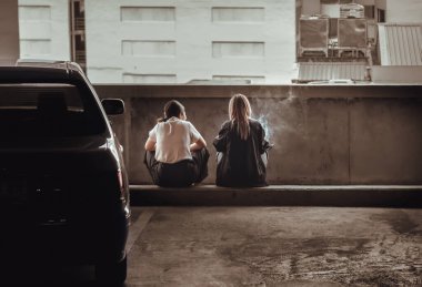 Bangkok, Tayland - 12 Ekim 2022: İki genç kadın, boş bir otoparkta beton durdurucunun üzerinde oturuyor ve sigara içiyor ya da havada asılı duruyor ya da elektronik sigara içiyor. Arka plan, kız molası, Karanlık ton, Seçici odak.