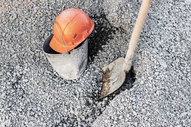 Betonu hazırlamak için moloz yığınına boyanmış bir kürek ve kovanın üzerinde duran turuncu bir miğfer.