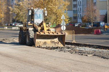 Ağır bir yükleyici toprağı kovayla taşır ve yolu derecelendirir. Sonbaharda modern bir şehrin merkezinde yol onarımı. Toprak taşımak için ağır inşaat makineleri
