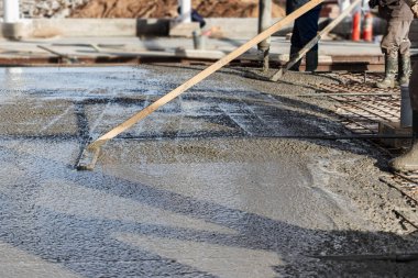 İnşaat işçisi endüstriyel bir atölyede beton döşemeyi yıkıyor. Bacakları betonun içinde. Yüzey somutlaşıyor. Monolitik güçlendirilmiş beton çalışmalar