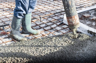 İnşaat işçisi endüstriyel bir atölyede beton döşemeyi yıkıyor. Bacakları betonun içinde. Yüzey somutlaşıyor. Monolitik güçlendirilmiş beton çalışmalar