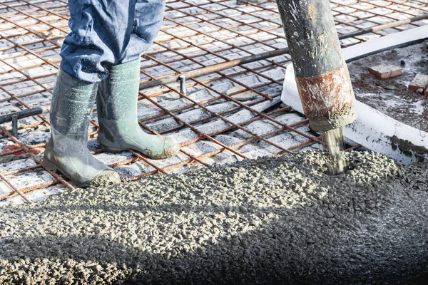 İnşaat işçisi endüstriyel bir atölyede beton döşemeyi yıkıyor. Bacakları betonun içinde. Yüzey somutlaşıyor. Monolitik güçlendirilmiş beton çalışmalar