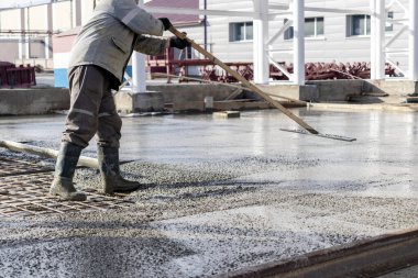 İnşaat işçisi endüstriyel bir atölyede beton döşemeyi yıkıyor. Bacakları betonun içinde. Yüzey somutlaşıyor. Monolitik güçlendirilmiş beton çalışmalar