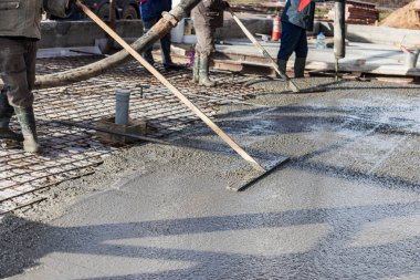 İnşaat işçisi endüstriyel bir atölyede beton döşemeyi yıkıyor. Bacakları betonun içinde. Yüzey somutlaşıyor. Monolitik güçlendirilmiş beton çalışmalar
