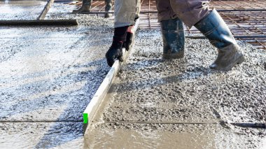 İnşaat işçisi endüstriyel bir atölyede beton döşemeyi yıkıyor. Bacakları betonun içinde. Yüzey somutlaşıyor. Monolitik güçlendirilmiş beton çalışmalar