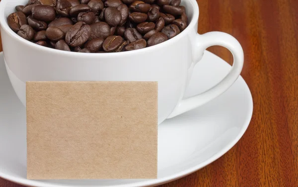 Xícara branca de grãos de café com cartão para texto em mesa de madeira — Fotografia de Stock