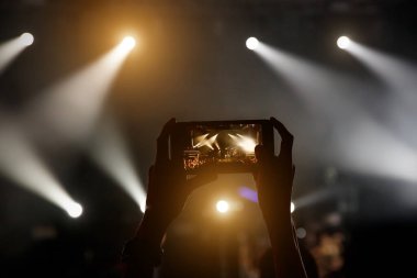Konser programındaki kadınların elindeki akıllı telefonun silueti, sahne ışığı.