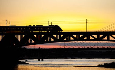 Tren gün batımında nehrin üzerindeki köprüden geçiyor.