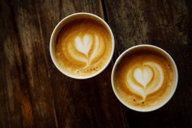 Two disposable cups of coffee with the heart on foam on a wooden table