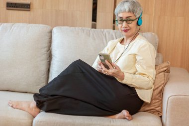 Kanepede oturup akıllı telefondan müzik dinleyen mutlu Asyalı kadın evde dinleniyor. 