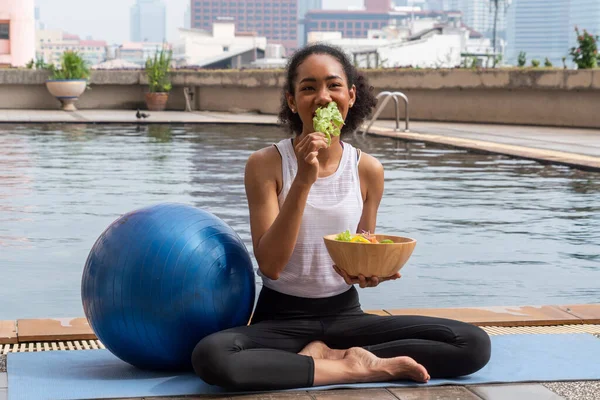 black woman sitting by the pool on a yoga mat having a blue yoga ball, finishes her workout, eats vegetable salad, healthy food and loses weight. Wellness and healthy concept