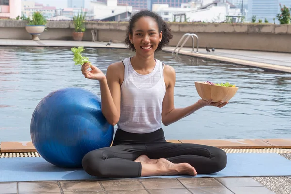 black woman sitting by the pool on a yoga mat having a blue yoga ball, finishes her workout, eating vegetable salad, healthy food and loses weight. Wellness and healthy concept