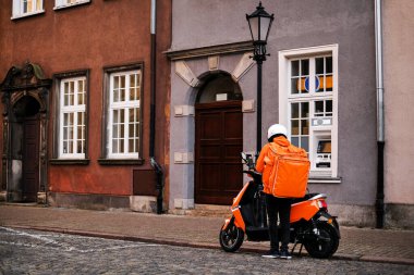  Motosikletli parlak üniformalı teslimatçı bir restorandan müşteriye teslimat siparişi almış. Teslimatçı telefonundaki dümenciye bakıyor.