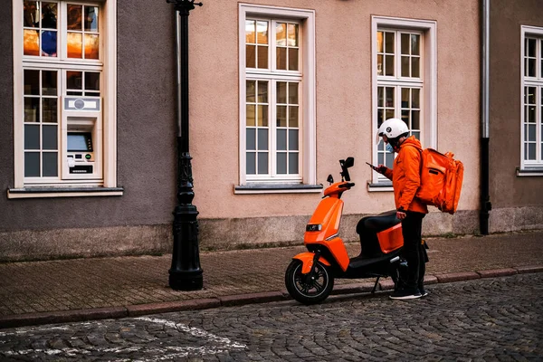  Motosikletli parlak üniformalı teslimatçı bir restorandan müşteriye teslimat siparişi almış. Teslimatçı telefonundaki dümenciye bakıyor.