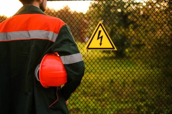 an engineer in uniform with a safety helmet stands at the entrance to a power plant with a danger sign on the fence