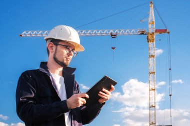 a builder looks at a tablet against the background of a house under construction with a crane