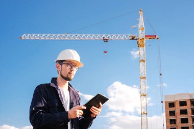 a builder looks at a tablet against the background of a house under construction with a crane