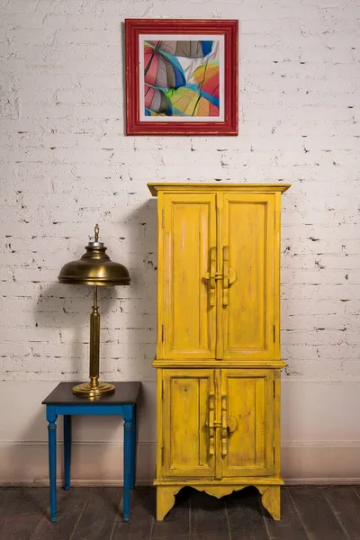 Armario amarillo vintage, pintura, lámpara de mesa de latón y mesa vintage en suelo de parquet de madera oscura y pared de ladrillos blancos — Foto de Stock