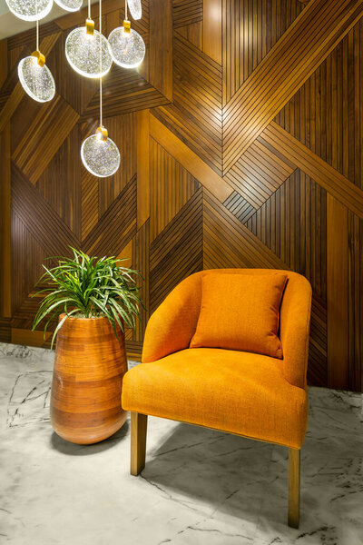 Modern orange armchair, tall rounded wooden planter with green bushes, tall glass chandelier, and wood cladding wall