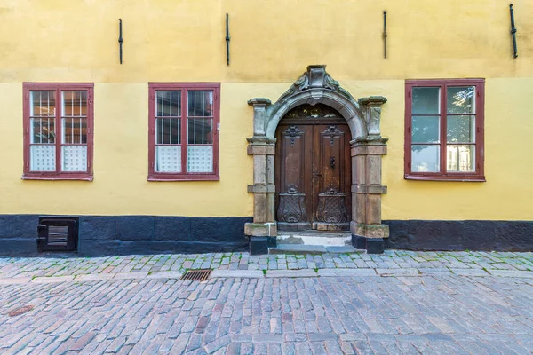 Vintage decorated wooden door framed by stone engraved frame, mediating three wooden and wrought iron windows in a grunge yellow wall at street with cobblestone floor, Old town, Stockholm, Sweden