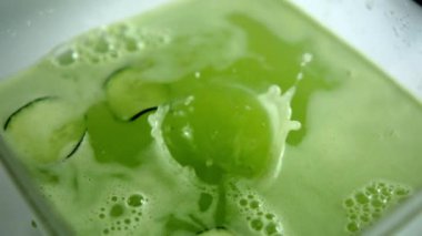 Salatalık dilimi cam kabın içindeki ferahlatıcı yeşil içeceğe dökülüyor.