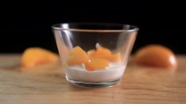 Şeftali dilimleri cam bardağa dökülüyor ve ahşap yüzeyde yoğurt var.