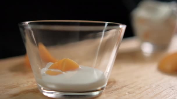 Plasterki brzoskwiń wpadające do szklanej filiżanki z jogurtem nad drewnianą powierzchnią — Wideo stockowe