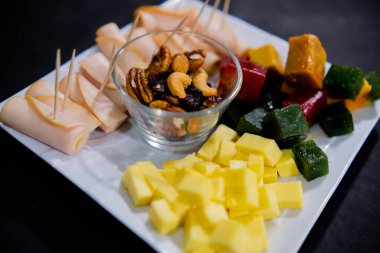 Jambonlu rulo, doğranmış meyve ezmesi, peynir küpleri ve kare beyaz tabakta fındıklar.