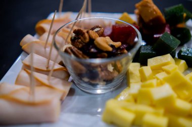 Jambonlu rulo, doğranmış meyve ezmesi, peynir küpleri ve kare beyaz tabakta fındıklar.