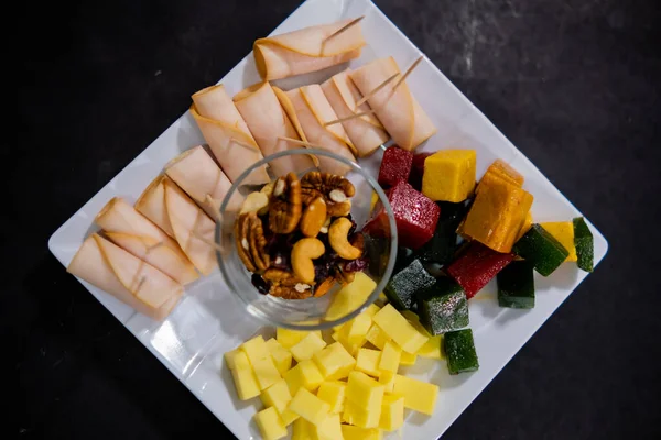 Ham rolls, diced fruit paste, cheese cubes, and nuts on square white plate