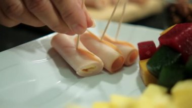 Jambon rulolarını dilimlenmiş meyve ezmesi ve peynir küpleriyle bir tabağa koyan eller.
