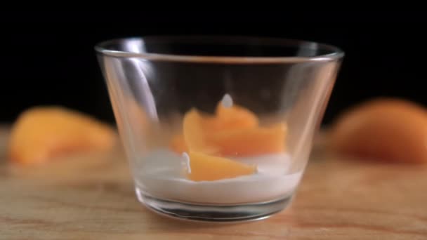 Fatias de pêssego caindo em copo de vidro com iogurte acima da superfície de madeira — Vídeo de Stock