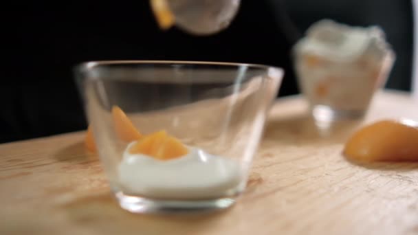 Perzikschijfjes die in glazen beker vallen met yoghurt boven houten oppervlak — Stockvideo