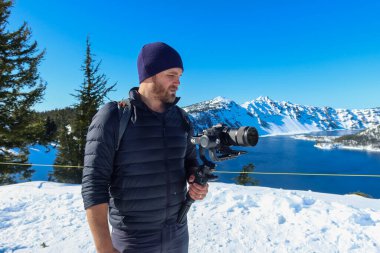 Sakallı adam arka plan olarak güzel karlı sıradağları olan bir kamera tutuyor.