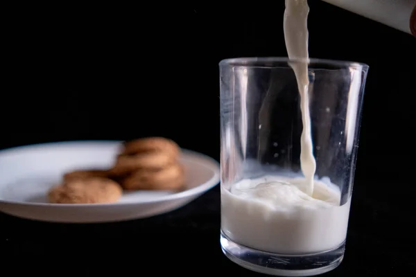 Verter la leche en un vaso junto al plato de galletas con fondo negro —  Fotos de Stock