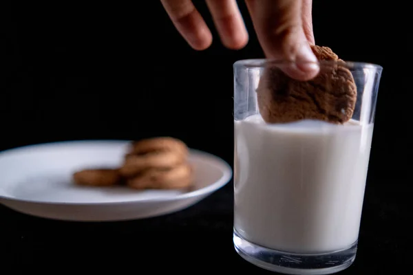 Mano mojando una galleta en vaso de leche con fondo negro — Foto de Stock