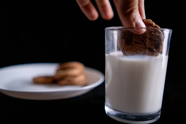 Рука макая печенье в стакан молока с черным фоном — стоковое фото