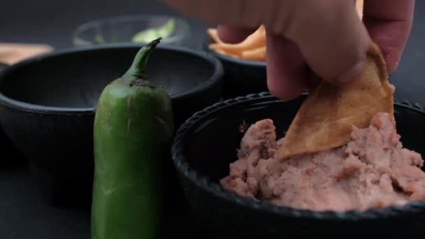 Hand dipping tortilla chip in refried beans in bowl behind a chili pepper — Stock Video