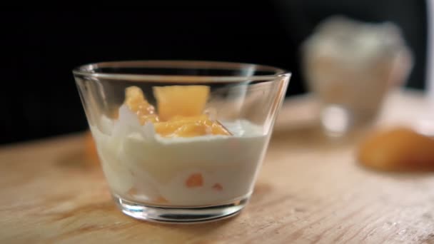Персиковые ломтики падают в стеклянную чашку с йогуртом над деревянной поверхностью — стоковое видео