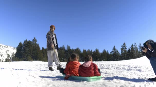 Mann fotografiert Kinder auf Schlitten mit blauem Himmel und Kiefern als Hintergrund — Stockvideo