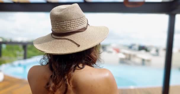 Mujer morena con sombrero lanzando su pelo hacia atrás con piscina borrosa como fondo — Vídeos de Stock