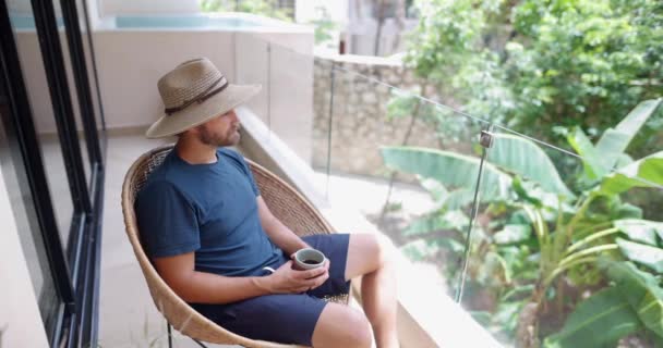 Человек, сидящий на тростниковом стуле на балконе и пьющий кофе — стоковое видео