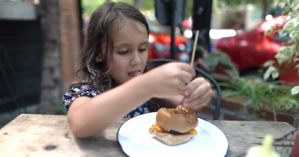 Linda niña pegando gran palillo de dientes en sabrosa hamburguesa — Vídeo de stock