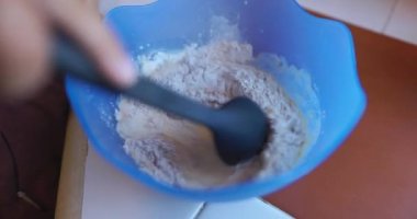 Büyük kaşıkla yumurta, un ve sütü karıştıran kişi mavi kasede.