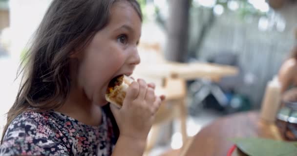 Милая маленькая девочка за деревянным столом ест буррито с размытым фоном — стоковое видео