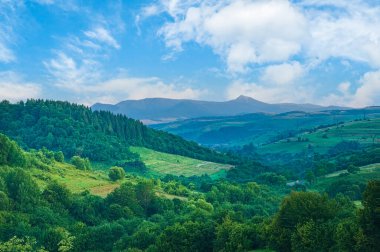 Dağ manzarası. Tepede çimenli tarlaları olan güzel bir dağ manzarası. Güneşli bir günde, uzak vadide, bulutlu bir köyün kırsal manzarası..