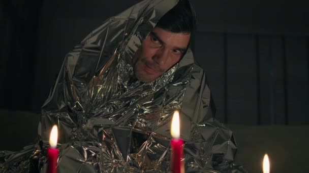 Strange man covered by a foil blanket looking on candles — Stock Video