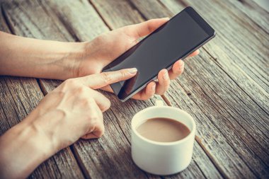 Ahşap bir masanın ve bir fincan kahvenin üzerinde ellerinde akıllı bir telefon..