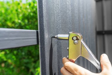 Bir adamın eli perçin tutuyor. Bir işçi, çitin üzerindeki profilli bir çarşafa perçin takar..